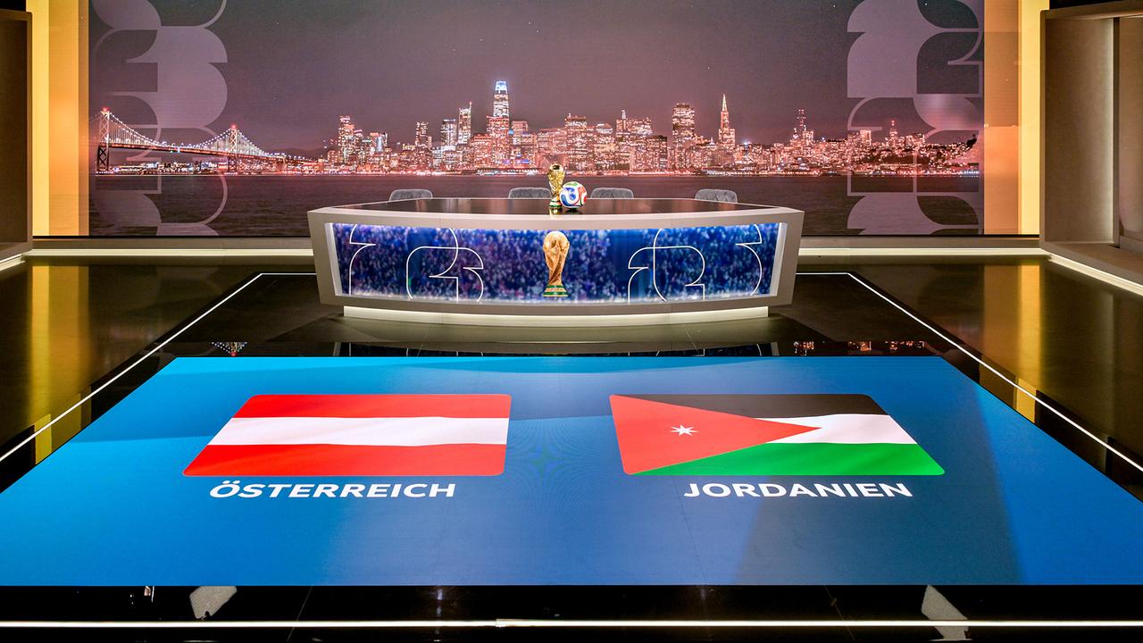 ORF-Fußball-WM-Studio mit einem Moderationstisch mit WM‑Pokal, dahinter eine nächtliche Stadtansicht und am Boden die Flaggen von Österreich und Jordanien.