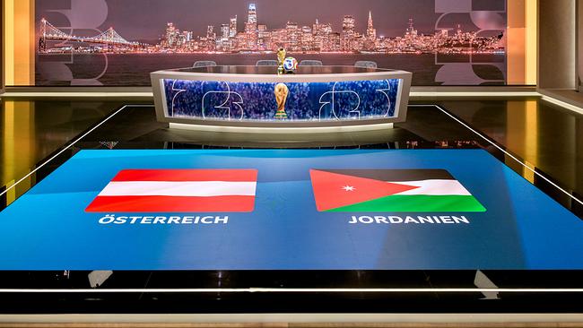 © ORF/Thomas Ramstorfer ORF-Fußball-WM-Studio mit einem Moderationstisch mit WM‑Pokal, dahinter eine nächtliche Stadtansicht und am Boden die Flaggen von Österreich und Jordanien.