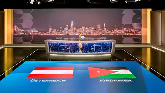 ORF-Fußball-WM-Studio mit einem Moderationstisch mit WM‑Pokal, dahinter eine nächtliche Stadtansicht und am Boden die Flaggen von Österreich und Jordanien.