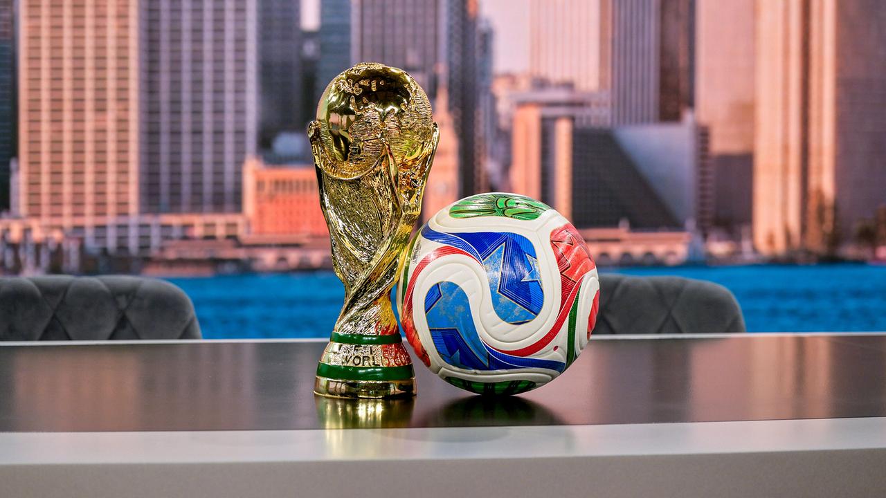 Der Fußball‑WM‑Pokal steht neben einem bunten Spielball auf einem Tisch vor einer urbanen Skyline‑Kulisse im neuen ORF WM-Studio