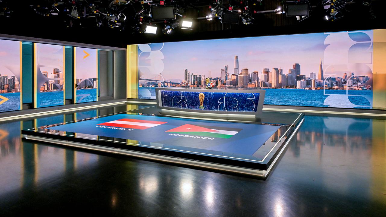 ORF-Fußball-WM-Studio mit einem Moderationstisch mit WM‑Pokal, dahinter eine nächtliche Stadtansicht und am Boden die Flaggen von Österreich und Jordanien.