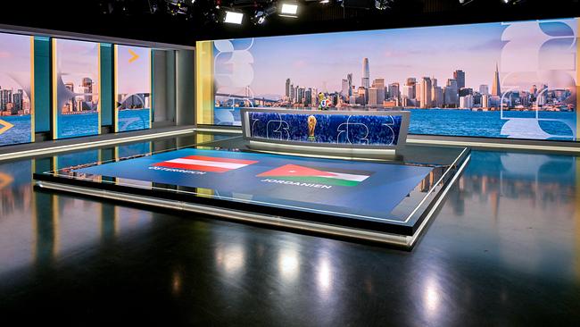 © ORF/Thomas Ramstorfer ORF-Fußball-WM-Studio mit einem Moderationstisch mit WM‑Pokal, dahinter eine nächtliche Stadtansicht und am Boden die Flaggen von Österreich und Jordanien.