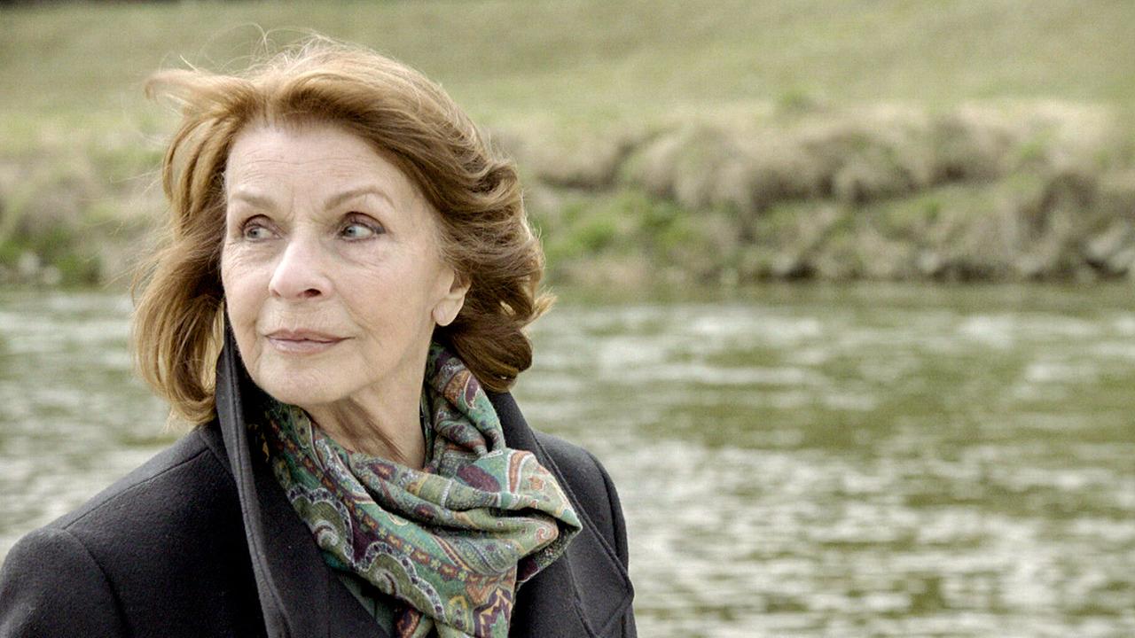 Senta Berger spaziert mit Schal und Mantel am Fluss.