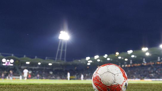 Ein rot-weiß-roter Fußball der Bundesliga liegt in der Nacht im Stadion am Rasen und im Hintergrund brennt das Flutlicht.