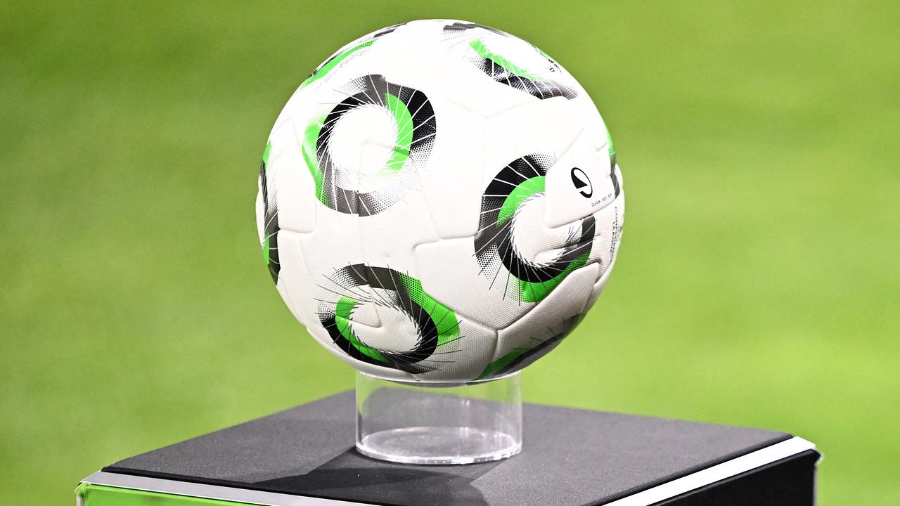 Ein weißer Fußball mit schwarz-grünem Muster liegt auf einem transparenten Sockel über einer schwarz-grünen Box auf einem Fußballplatz.