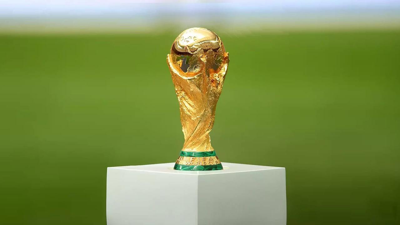 Der goldene FIFA‑WM‑Pokal steht gut ausgeleuchtet auf einem weißen Podest vor einem grünen Spielfeld.