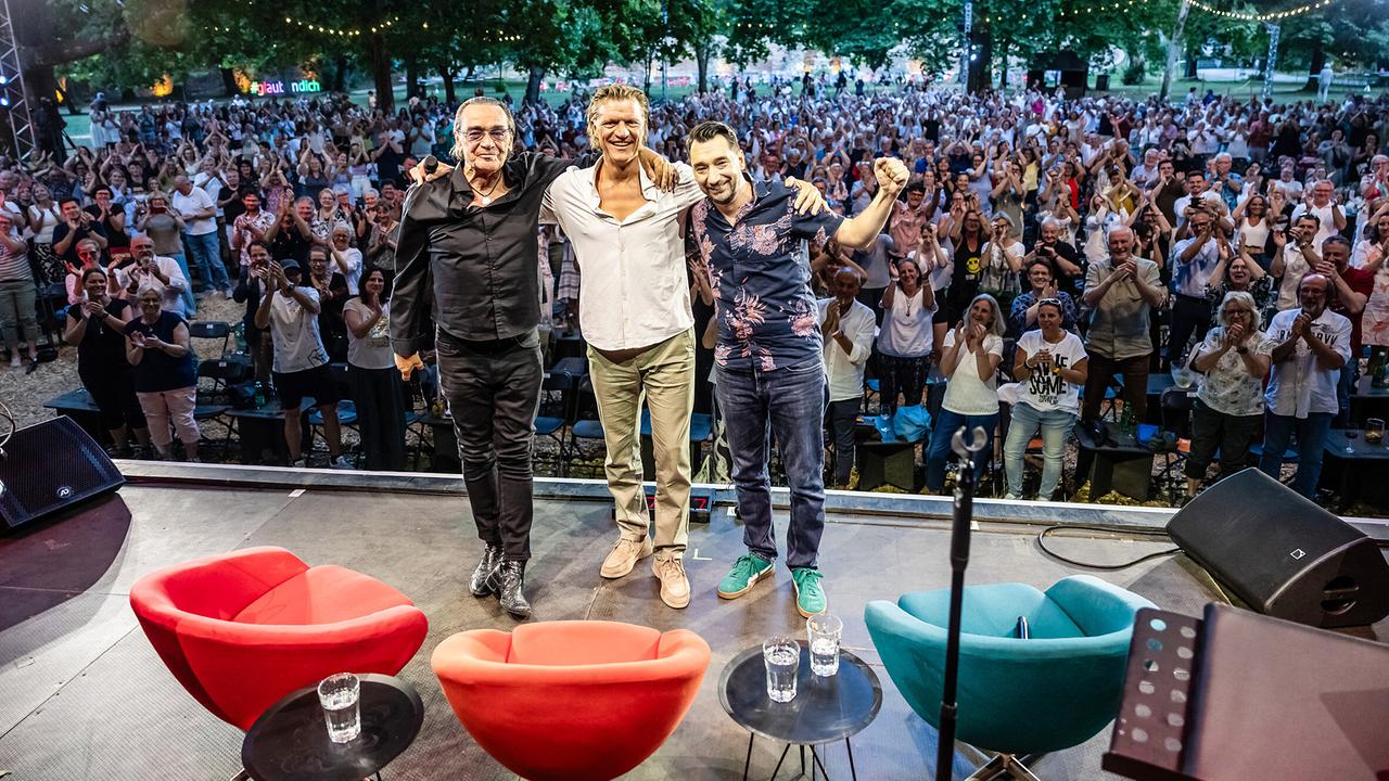 Christian Kolonovits, Gery Seidl und Bernhard Egger stehen auf der Open-Air-Bühne vor dem Publikum im Wiener „Theater im Park“ bei „Auf dem Roten Stuhl“.
