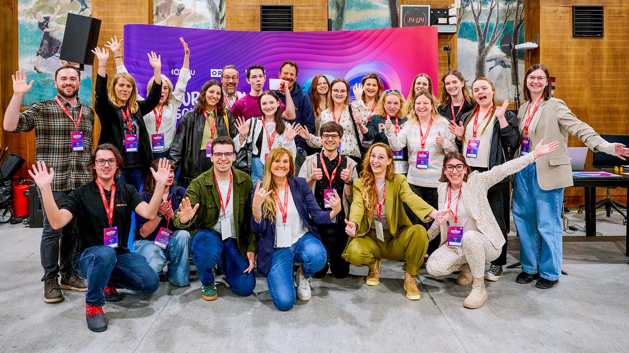 © ORF/Klaus Titzer A large group of participants at the volunteers casting cheerfully pose with raised hands in front of a colorful event stage with name badges around their necks.
