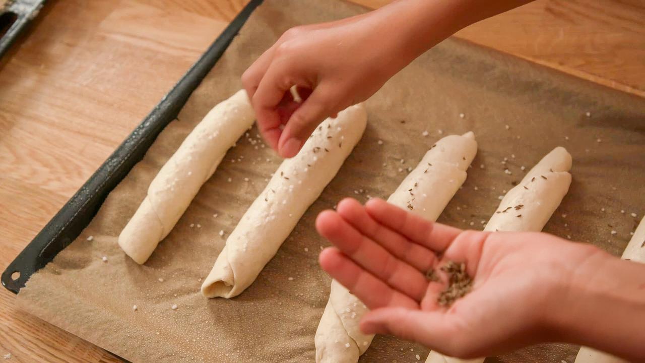 Jemand bestreut längliche Salzstangerln auf einem mit Backpapier belegten Blech mit Salz und Kümmel.