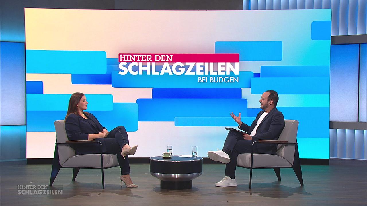 Barbara Karlich sitzt gegenüber von Patrick Budgen an einem Coachtisch vor einer großen, farbigen Studiografik mit dem Schriftzug "Hinter den Schlagzeilen bei Budgen"