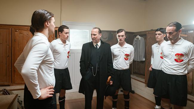 In einer historischen Umkleidekabine stehen mehrere Fußballspieler in alten Trikots um Trainer Hugo Meisl im Anzug, der eine Ansprache hält.