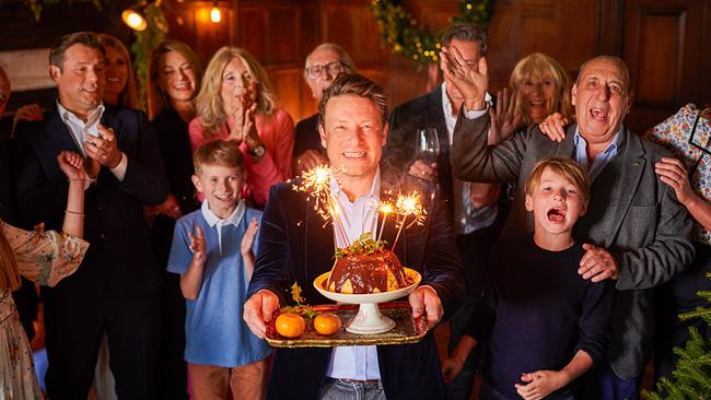 Jamie Oliver hält einen Bananen-Panettone-Pudding mit Schokolade-Einschlüssen und Wunderkerze. Rund um ihn sind seine Familie und Freunde versammelt