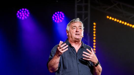 Andreas Vitásek  steht gestikulierend auf einer beleuchteten Bühne, während farbige Scheinwerfer im Hintergrund leuchten.