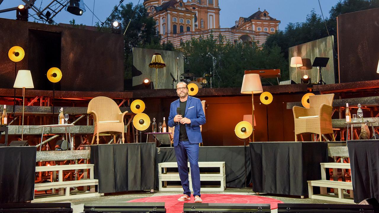 © ORF/Hubert Mican Gerald Fleischhacker steht im Rahmen von "Kabarett unter Sternen: Die Tafelrunde Open Air" bei Dämmerung auf der Bühne der Wachauarena vor dem Stift Melk