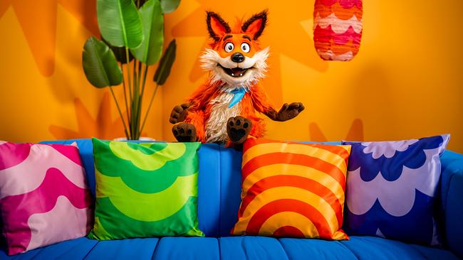 © ORF/Steiner Familyentertainment/Florian Falb Fuchsi, eine fröhliche Fuchs‑Handpuppe, lehnt an einem Tisch in einem bunt gestalteten Studio mit Regalen und Pflanzen im Hintergrund.