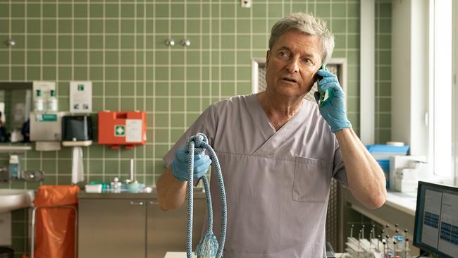 Alfred Dorfer als Pathologe Dr. Tom Wippler in medizinischer Arbeitskleidung und Handschuhen telefoniert in einem klinischen Raum und hält dabei ein Seil in der Hand.