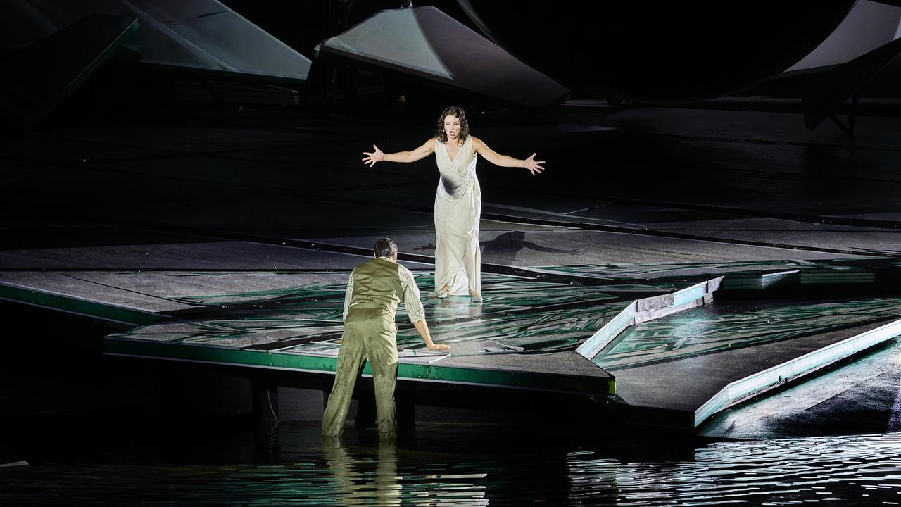 Marjukka Tepponen als Violetta im weißen Kleid breitet die Arme aus und singt einem Mann entgegen, der vom Wasser aus zur Bühnenkante hochklettert.