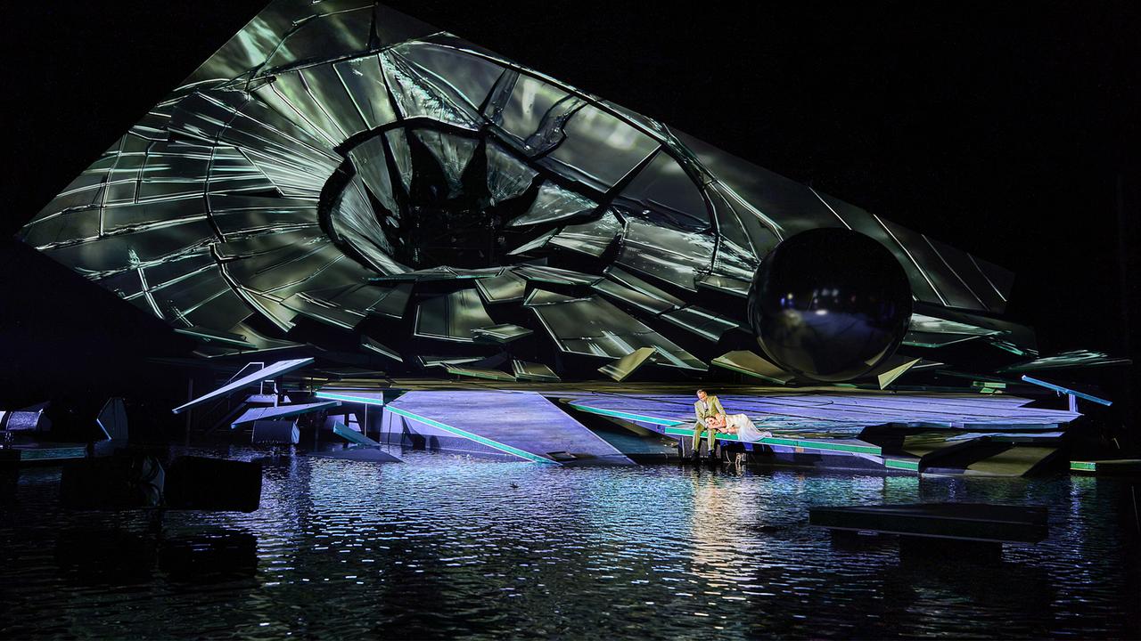 Eine große, wie zerbrochen wirkende Scheibe dominiert die nächtliche Seebühne der Bregenzer Festspiele, auf deren Rand ein Paar sitzt.