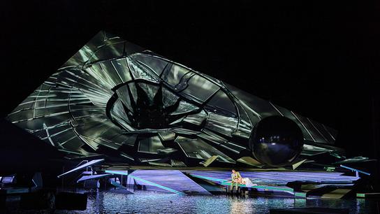 Eine große, wie zerbrochen wirkende Scheibe dominiert die nächtliche Seebühne der Bregenzer Festspiele, auf deren Rand ein Paar sitzt.