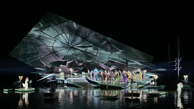© ORF/Bregenzer Festspiele/Anja Köhler Eine große Gruppe von Sänger:innen steht auf der hell erleuchteten Seebühne in Bregenz vor einer monumentalen, zerbrochen wirkenden Scheiben-Kulisse, während einzelne Figuren seitlich und auf Plattformen im Wasser agieren.