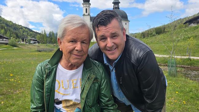 Marc Pircher und G.G. Anderson posieren auf einer grünen Wiese, im Hintergrund sind Berge und eine Kirche mit Zwiebeltürmen unter blauem Himmel zu sehen.