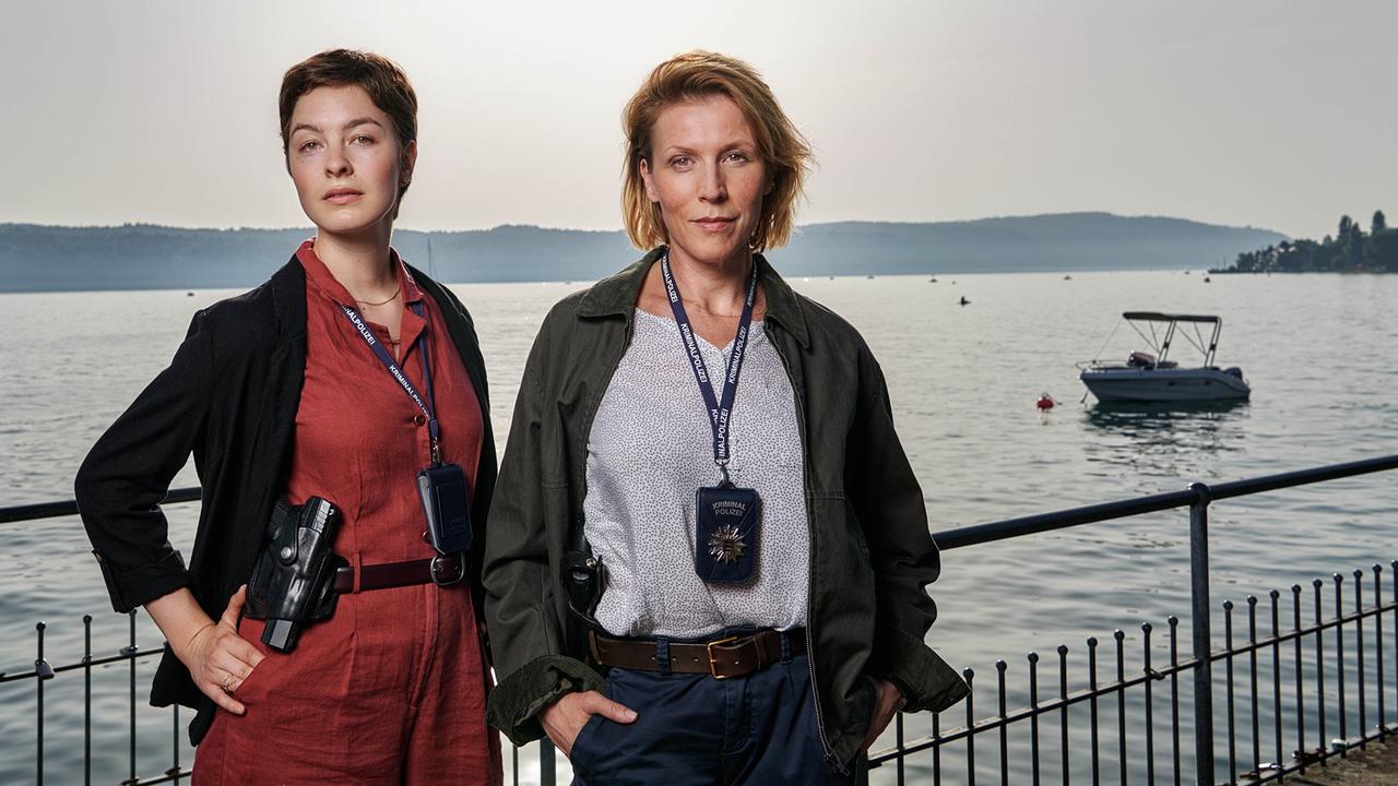 © ORF/ZDF/Patrick Pfeiffer Konstanz Maria Wördemann und Franziska Weisz mit Ausweisen und Funkgeräten stehen an einer Uferpromenade vor dem Bodensee, im Hintergrund sind Boote, Wasser und eine hügelige Landschaft zu sehen.