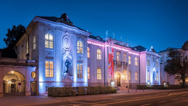 © ORF/Landesstudio Salzburg/Christian Schneider Ansicht der Stiftung Mozarteum bei Abenddämmerung, dessen Fassade in warmem Gelb und kühlem Blau angestrahlt wird.