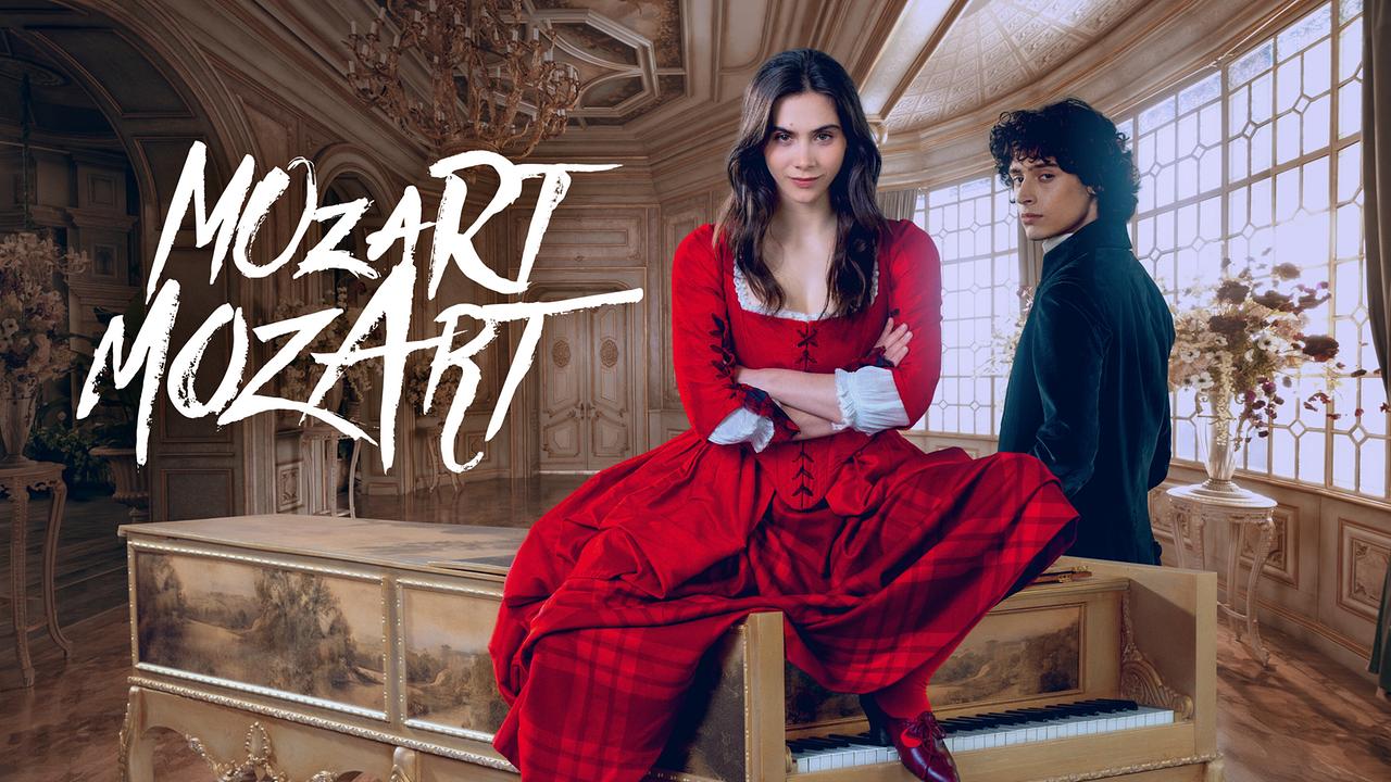 Havana Joy als Maria Anna Mozart in rotem Kleid sitzt auf einem Cembalo, während Eren M. Güvercin als Wolfgang Amadeus Mozart in dunkler Kleidung danebensteht, vor dem Schriftzug „Mozart Mozart“ in einem eleganten Raum.