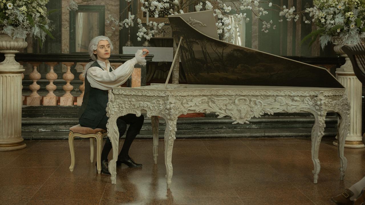 © ORF/WDR/Story House Pictures/Armands Virbulis Eren M. Güvercin als Wolfgang Amadeus Mozart in historischer Kleidung sitzt an einem reich verzierten Cembalo in einem opulent dekorierten Raum mit blühenden Zweigen.