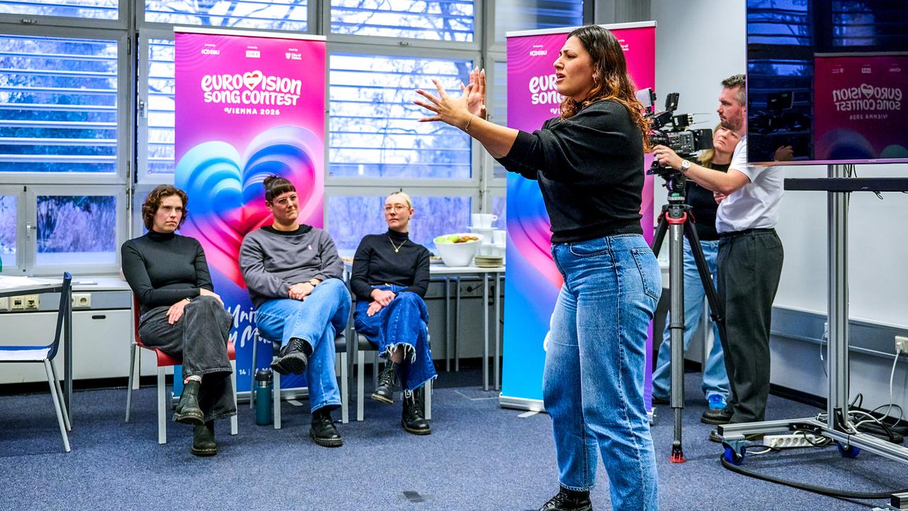© ORF/Klaus Titzer Carina Kilinc, a casting participant, gestures energetically toward a small seated audience while a camera operator films, with brightly colored Eurovision Song Contest Vienna banners visible behind them.