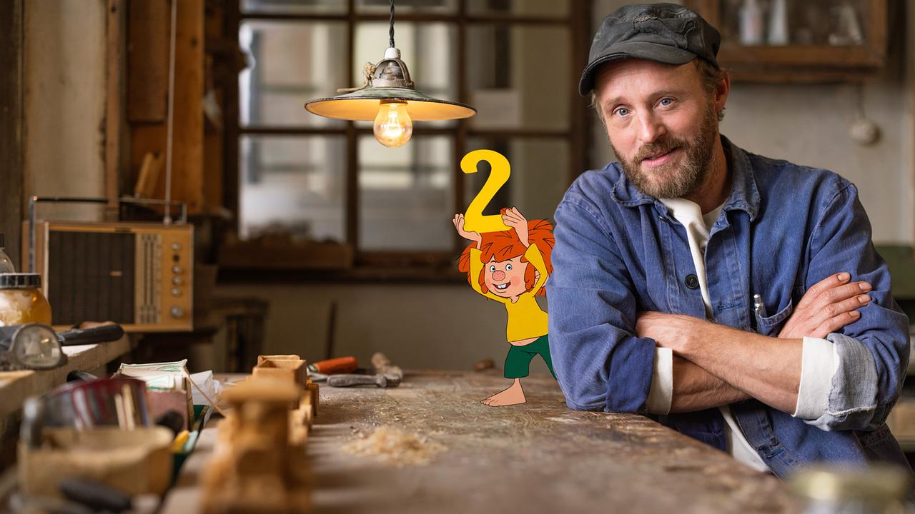 Pumuckl hält fröhlich eine große gelbe „2“ hoch und steht auf einer Werkbank neben Florian Eder in der Werkstatt.