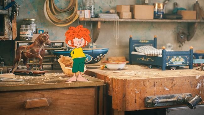 Pumuckl steht in der Schreinerwerkstatt auf einer Werkbank zwischen einem Holzpferd, einem kleinen Holzbett und Werkzeugen.