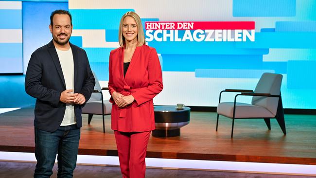 Patrick Budgen und Mariella Gittler stehen in einem modernen TV‑Studio vor einer großen Bildschirmwand mit dem Titel „Hinter den Schlagzeilen“, umgeben von Sitzmöbeln und Studiolicht.
