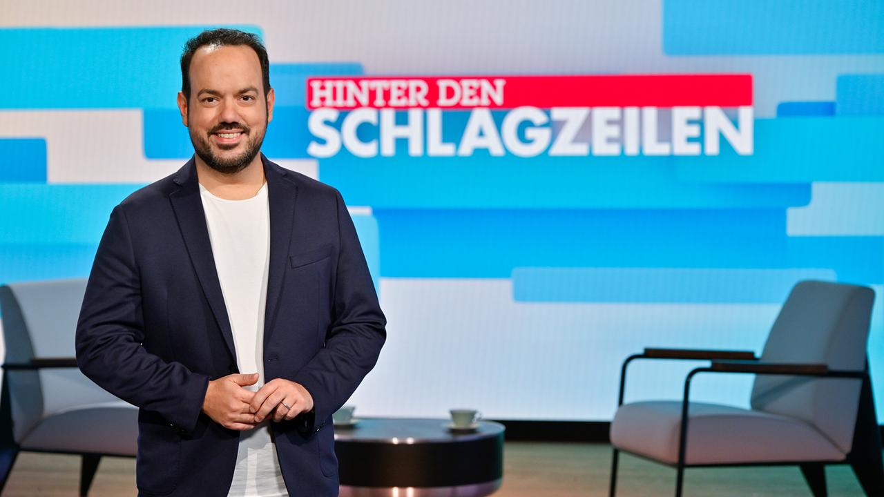 Patrick Budgen steht in einem modernen TV‑Studio vor einer großen Bildschirmwand mit dem Titel „Hinter den Schlagzeilen“, umgeben von Sitzmöbeln und Studiolicht.