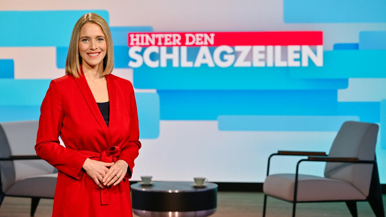 Mariella Gittler steht in einem modernen TV‑Studio vor einer großen Bildschirmwand mit dem Titel „Hinter den Schlagzeilen“, umgeben von Sitzmöbeln und Studiolicht.