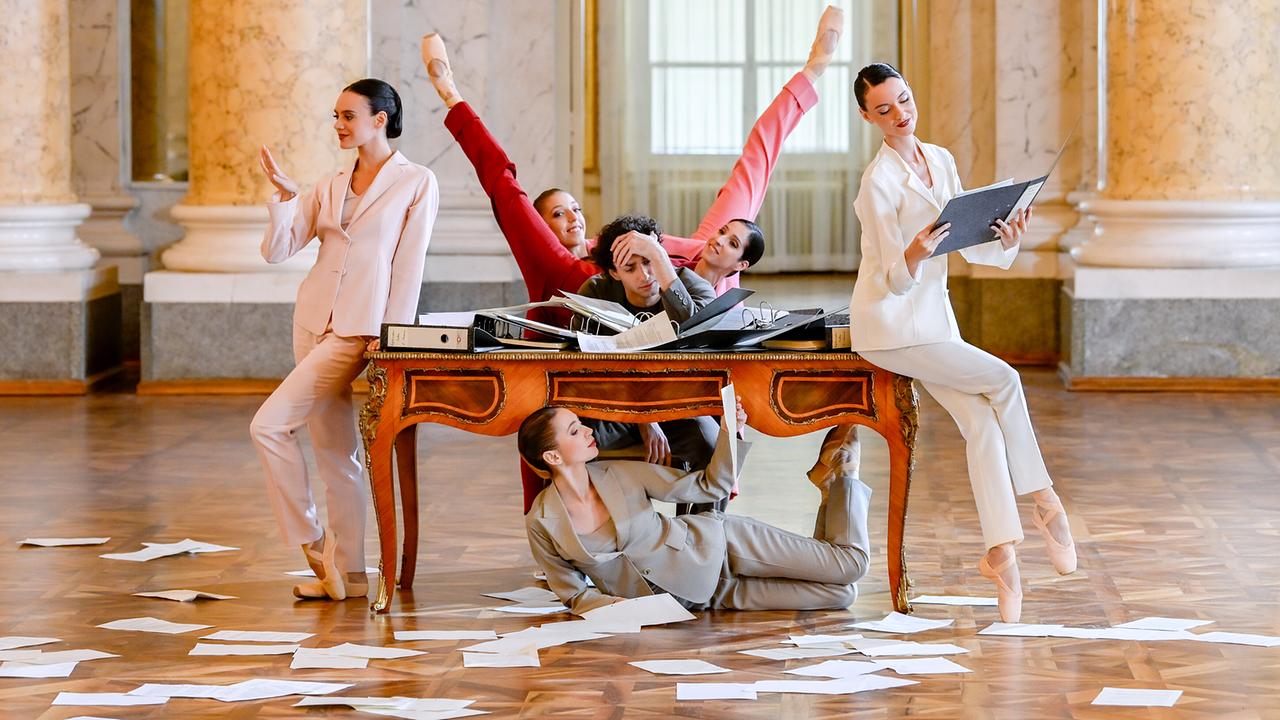 Die Tänzer:innen Natalya Butchko, Gaia Fredianelli, Anita Manolova, Alessandro Frola, Tatiana Mazniak und Laura Cislaghi in einer verspielten Szene rund um einen Schreibtisch mit verstreuten Papieren in einem prachtvollen Saal in Szene.