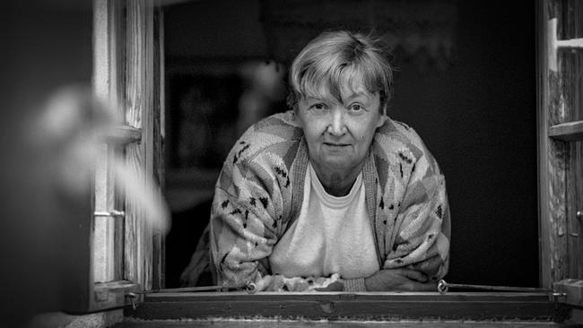 Das Schwarz-Weiß-Foto zeigt Christine Nöstlinger, die sich aus einem geöffneten Holzfenster lehnt und direkt in die Kamera blickt.