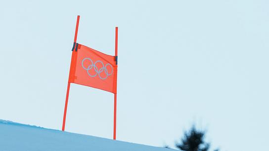 Ein rotes Tor mit olympischen Ringen steht im Schnee vor hellem Himmel.