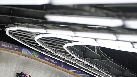 Der Rodler Wolfgang Kindl beim Training im Eiskanal, der im beleuchteten Cortina Sliding Centre eine Steilkurve einer Bahn mit „Milano Cortina 2026“-Beschriftung durchfährt.