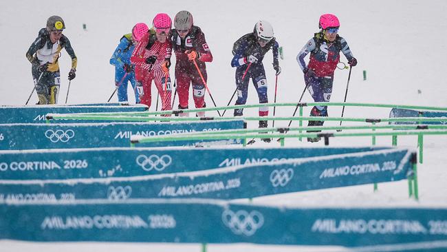 Sechs Sprint-Skibergsteigerinnen laufen im Schneefall über einen Kurs, vorbei an Absperrbändern mit „Milano Cortina 2026“ und den Olympischen Ringen im Skizentrum Stilfser Joch in Bormio.
