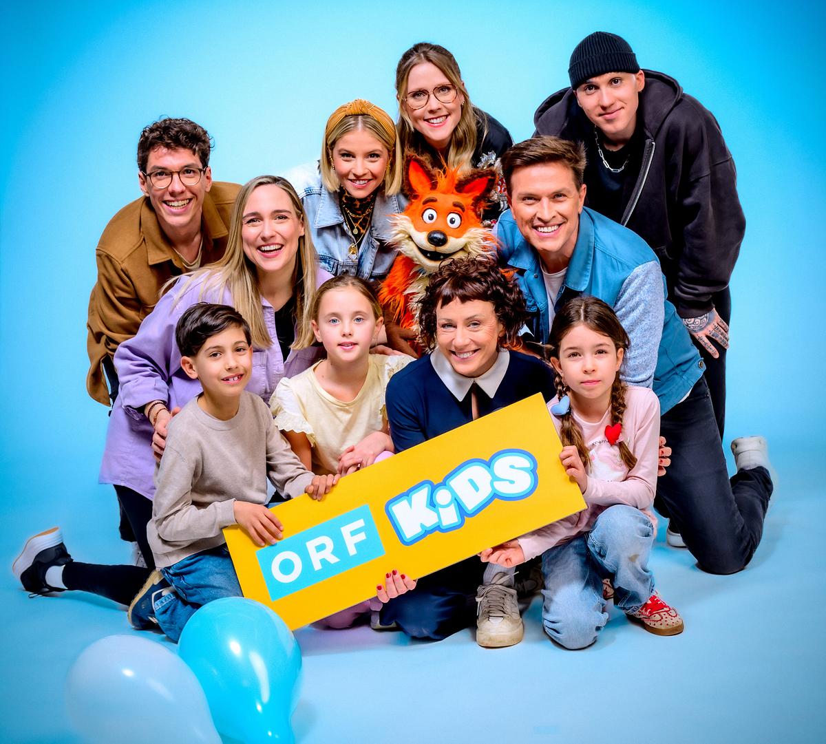 © ORF/Hans Leitner Christoph Hirschler, Melanie Flicker, Fanny Stapf, Lena Großmann, Esther Csapo, Robert Steiner, Stefan Gruber mit Fuchsi und Kindern posieren vor blauem Hintergrund und halten ein Schild mit der Aufschrift „ORF Kids“.