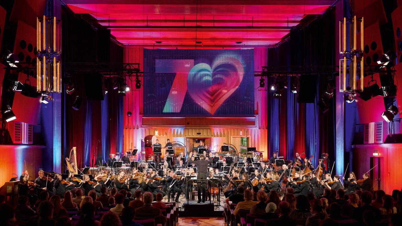 The image shows a large orchestra playing in the colorfully lit concert hall at the ORF RadioKulturhaus in front of an audience, while a glowing 70s heart motif is projected above the stage.