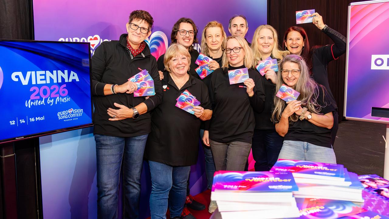 © ORF/Graph Art Line e.U. A group of people proudly hold up colorful Eurovision Vienna 2026 cards while posing together in front of a vibrant event backdrop.
