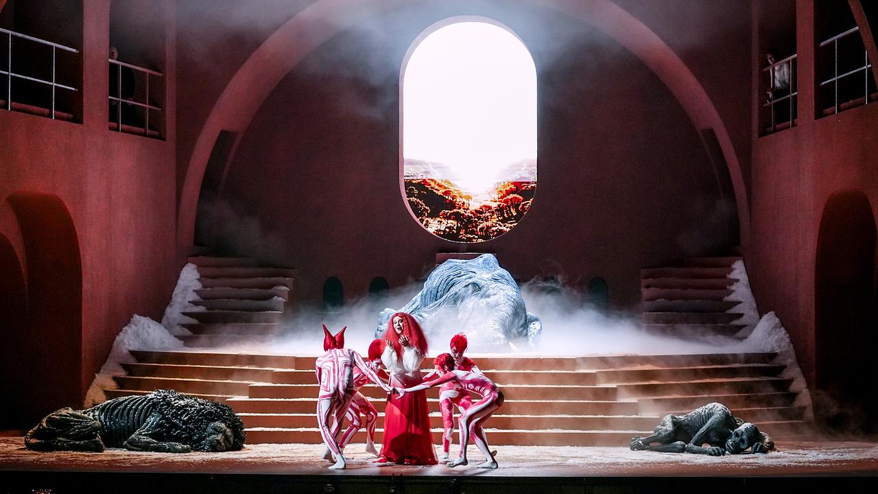 © ORF/Roman Zach-Kiesling Ariadne auf Naxos: Vor einer nebligen Treppe und einem hellen Fenster wird Araidne im roten Kleid von drei stilisiert kostümierten Figuren umringt.