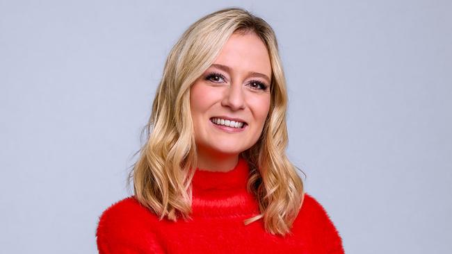 Tina Ritschl posiert mit verschränkten Armen in einem roten Rollkragenpullover und Jeans vor einem neutralen Hintergrund.