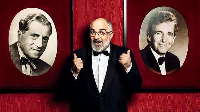 © ORF/Simpl/Jan Frankl Michael Niavarani im Smoking steht vor einer roten Wand zwischen zwei gerahmten Porträtfotos und hebt beide Daumen nach oben.