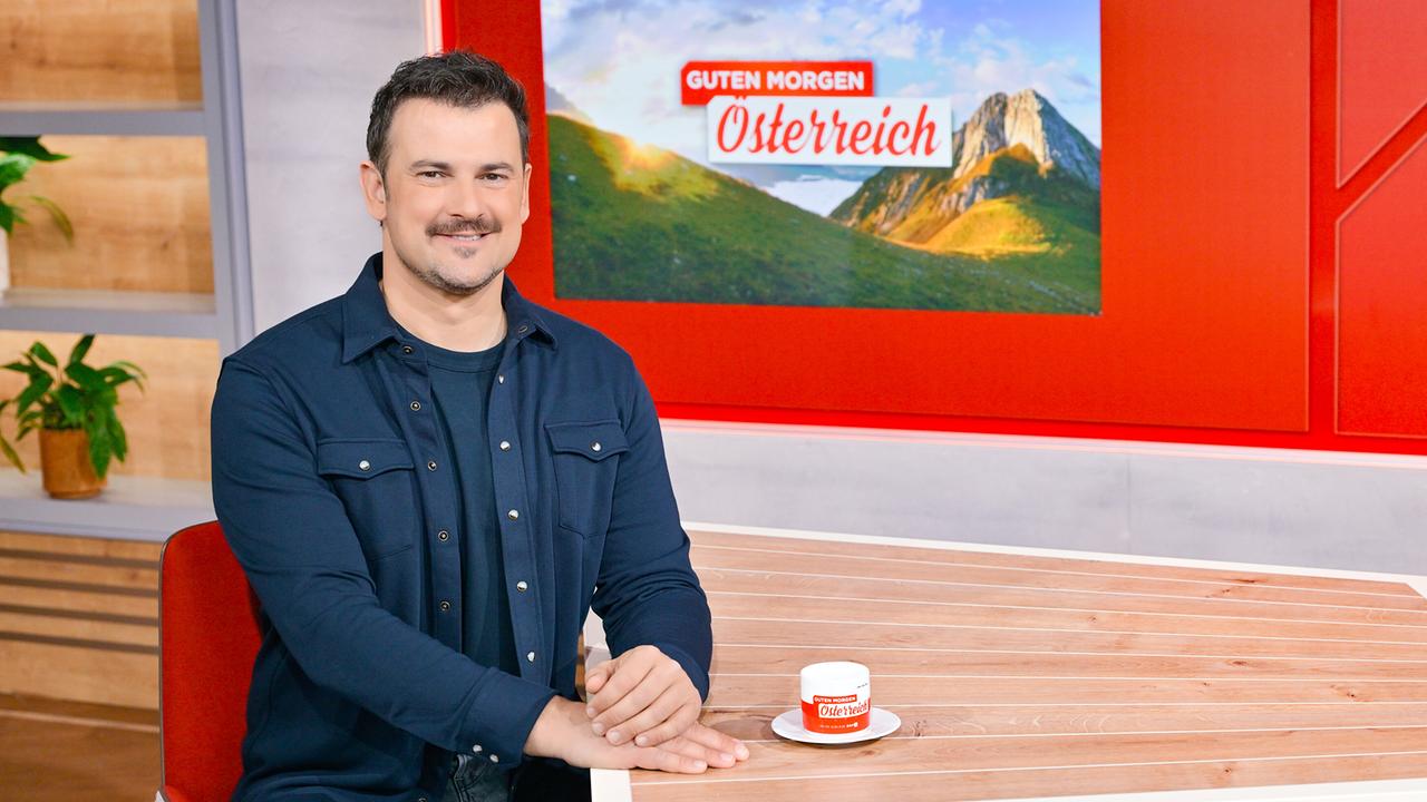 © ORF/Thomas Ramstorfer Sigi Fink sitzt an einem Holztisch im „Guten Morgen Österreich“-Studio, vor ihm steht eine kleine „Guten Morgen Österreich“-Tasse auf einer Untertasse.
