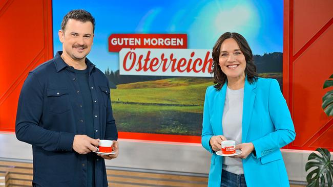 Sigi Fink und Eva Pölzl stehen im Studio von „Guten Morgen Österreich“ vor einem großen Bildschirm und halten jeweils eine Tasse.