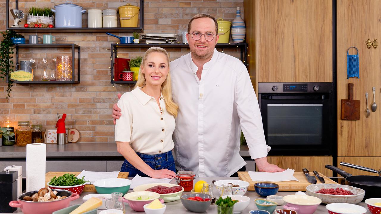 © ORF/Silvia Schneider GmbH/Juraj Melicher Silvia Schneider und Florian Unseld stehen in der gemütlich eingerichteten Studioküche hinter einer Arbeitsfläche mit zahlreichen vorbereiteten Zutaten.