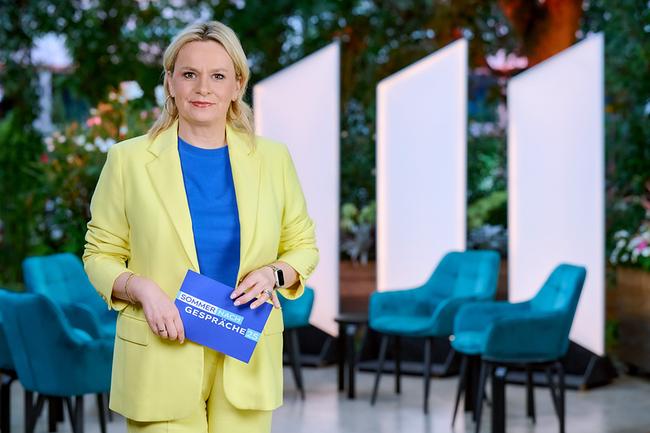 © ORF/Klaus Titzer Lou Lorenz-Dittlbacher steht in einem Garten‑Setting vor modernen Sesseln und Lichtpaneelen und hält eine Moderationskarte in der Hand.